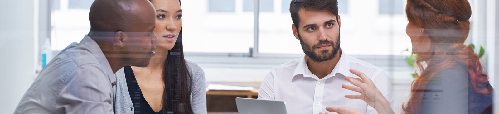 A small group of young colleagues sits around a laptop in an office setting, actively engaged in discussion.